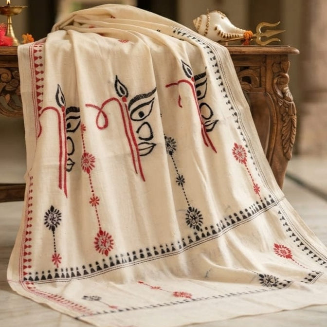An off-white cotton shawl with red and black embroidered motifs draped over a carved wooden table in a warm, softly lit indoor setting.